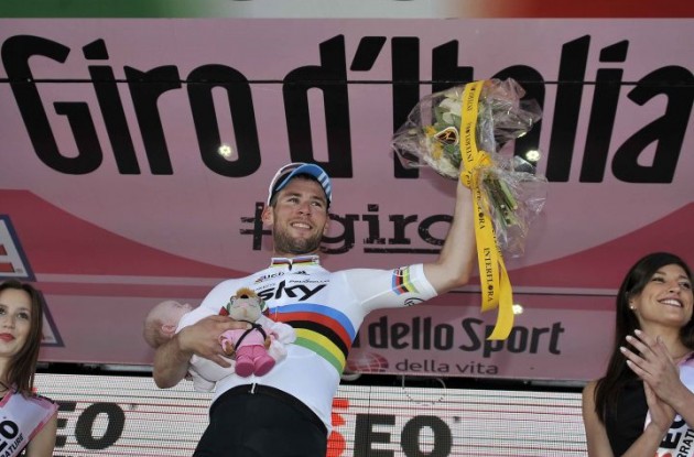 Mark Cavendish celebrates his stage win on the podium with his newborn child and the podium trophys.